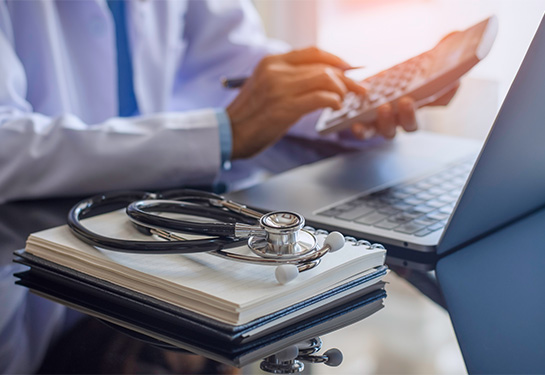 Person in white doctor&#x2019;s coat sits next to a stethoscope resting on a notebook 