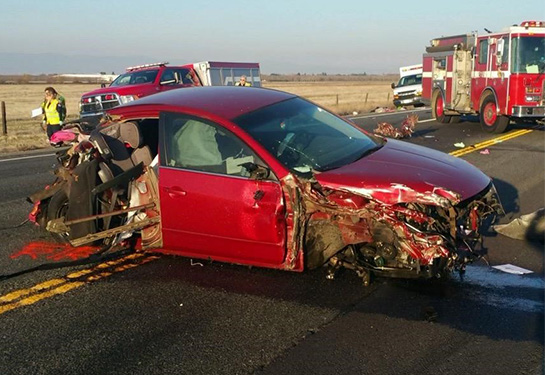 Red vehicle with damage to the front and back with a fire truck and ambulance in the background