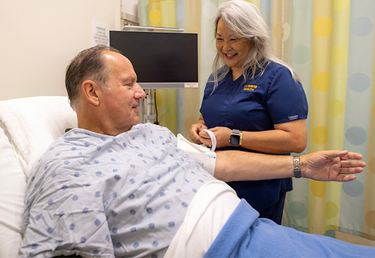 A man laying in a hospital bed extends his arm as a nurse puts a blood pressure cuff on his bicep.