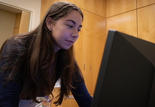 A woman with long brown hair and wearing a blue shirt leans over a desk to look at a computer monitor. 