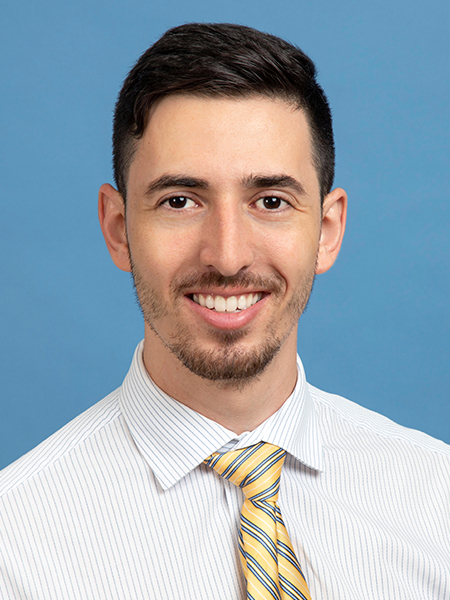 A man with dark hair, thin mustache and beard, wearing a white shirt and yellow tie