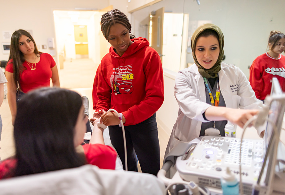 A student moves a device on the wrist of another student while a doctor points to a screen.