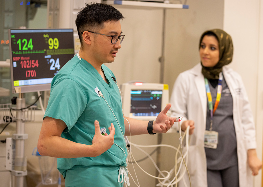 A man in green scrubs stands in front of a medical monitor and a woman in a white coat stands nearby.