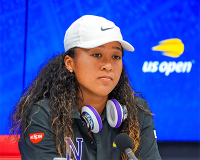 Naomi Osaka in a press conference with a headphone around her neck and a white Nike cap on her head