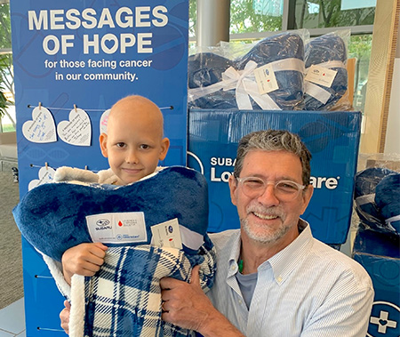 Bald young boy hugs navy-blue pillow blanket while standing next to man in a light-blue shirt who is kneeling.