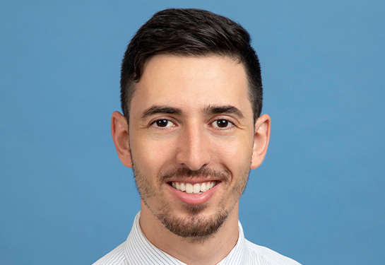 A man with dark hair, thin mustache and beard, wearing a white shirt and yellow tie