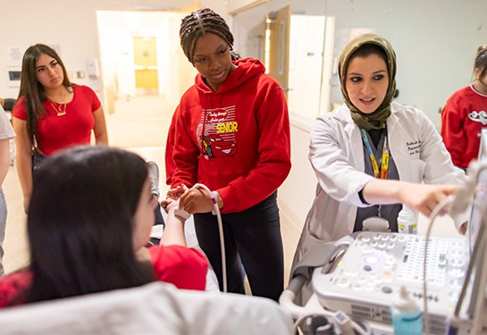 A student moves a device on the wrist of another student while a doctor points to a screen.