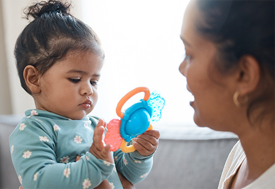 A toddler being held by her mother plays with a colorful plastic toy. 