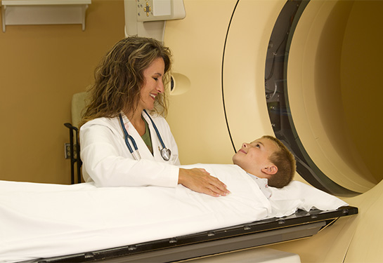 Child lays on gurney covered by white blanket as woman in white lab coat stands over him near circular CT machine. 