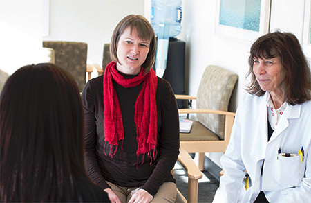 A woman in a white coat with dark hair and a woman with short hair and a black sweater with a red scarf talk to a person whose back is facing the camera.