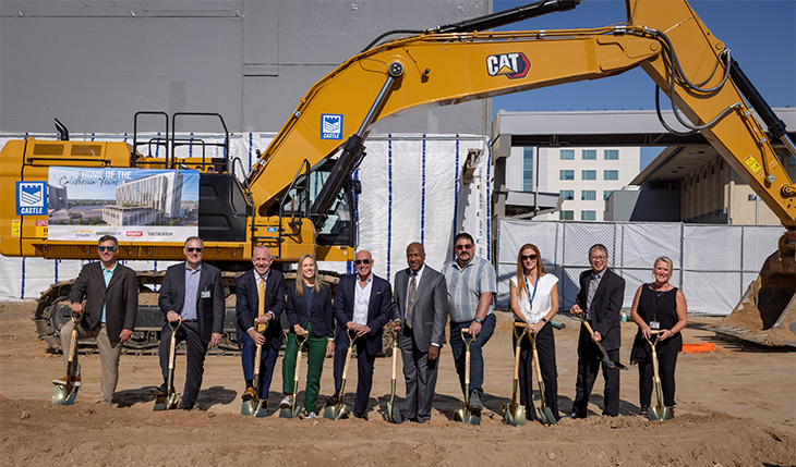 10 people standing in row holding gold shovels with a front loader behind them.