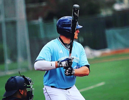 A man in blue helmet and light blue jersey grips a dark baseball bat awaiting the next pitch on a baseball field