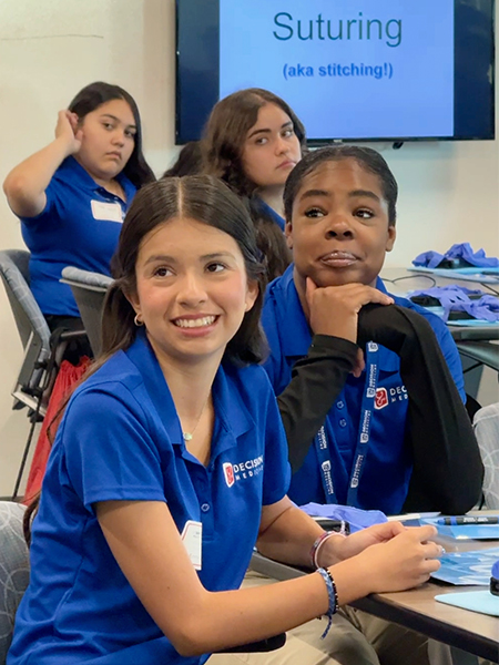 Four teenage girls in blue “Decision Medicine” polo shirts sit at a table as they receive instructions on how to suture a banana