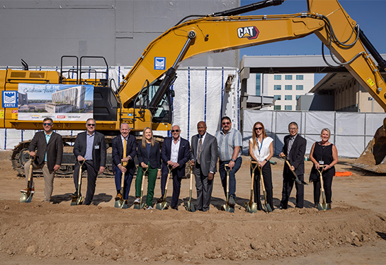 10 people standing in row holding gold shovels with a front loader behind them.