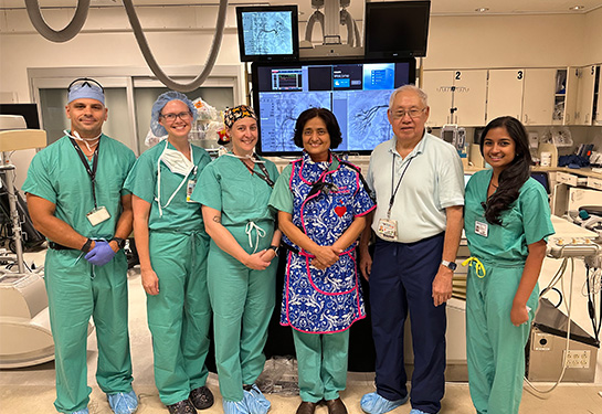 Six people standing in a row in an operating room with computer screens above their head.