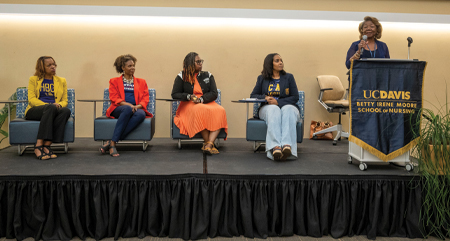 Four women sit on stage in chairs side by side while woman stands at podium holding microphone