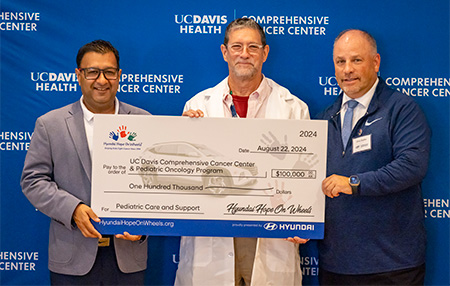 Three men hold oversized check for $100,000 made out to UC Davis Comprehensive Cancer Center & Pediatric Oncology Program for “pediatric care and support.”