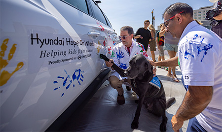 Black dog placing paw on car helped by two men, one in white coat painted in handprints, and one in white t-shirt painted in handprints.