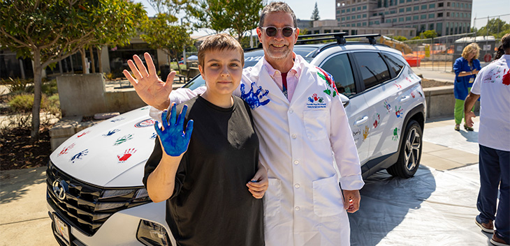 Girl with short hair and black t-shirt standing next to man in white coat as she holds up her palm painted blue.