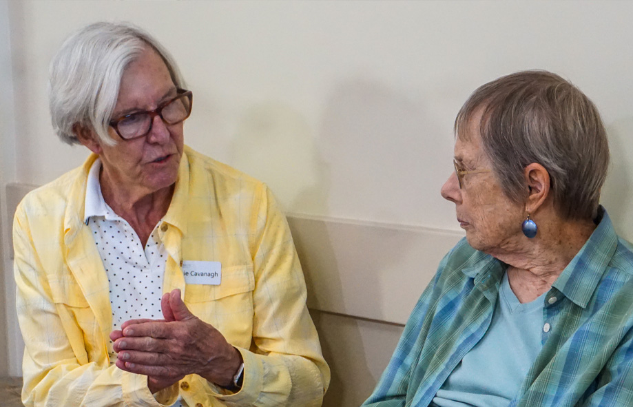 Two women face each other talking while seated against a wall