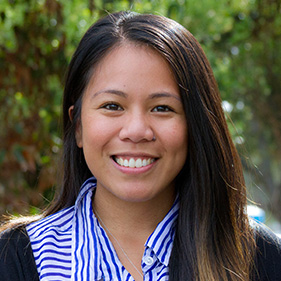 Catrina Calub, a woman with long dark hair wearing a blue and white shirt and black sweater sits outside smiling. 