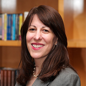 Julie Schweitzer, a woman with medium-length dark hair, wearing a gray suit, smiles in a library. 