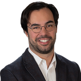 A man with dark hair, wearing a white shirt and dark suit coat smiles. 