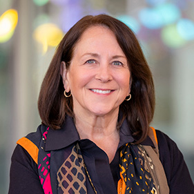 A smiling woman with brown hair, wearing a black shirt and black, orange and pink scarf.