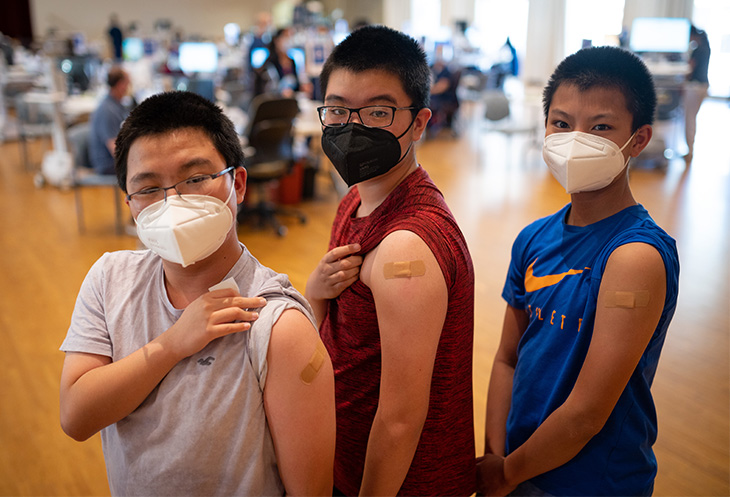 Three kids standing next to each other and showing their arms where they were vaccinated. They are all wearing masks.