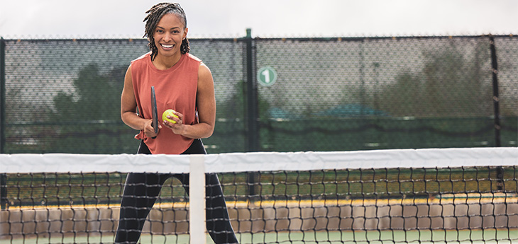 Adult wearing a pink tank top and black leggings stands on a pickleball court with a racket and ball.