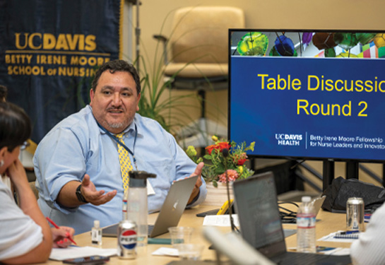 Man sits next to monitor that says “Table Discussion Round 2” and gestures with his right hand while talking to woman sitting next to him; Betty Irene