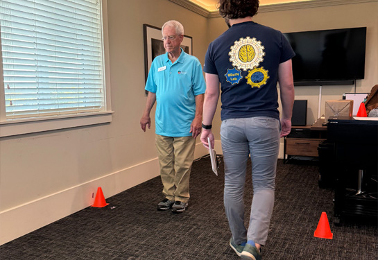  Man, left, stands between two orange cones on floor during balance assessment while researcher looks on