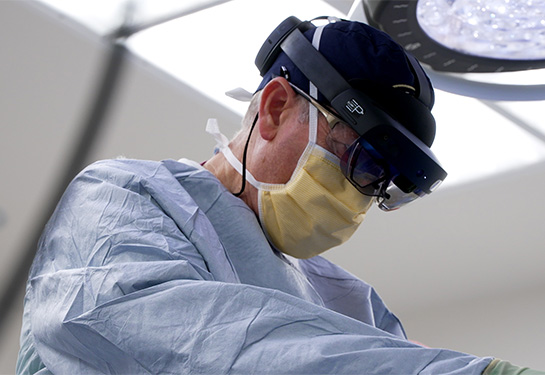 Surgeon wearing blue scrubs with bright lights behind him leans over patient wearing large black headset.