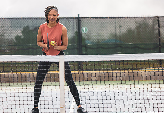 An adult wearing a pink tank top and black leggings stands on a pickleball court with a racket and ball. 