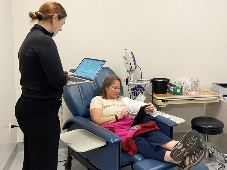 Clinical research coordinator wearing black top and pants is holding a laptop and explaining the study to a participant in a clinical trial. The participant is reading on a tablet.