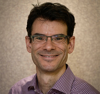 A man with brown hair, glasses and a maroon and white plaid shirt smiles