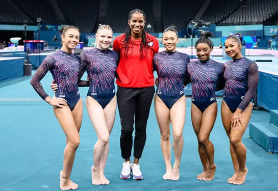 Marcia Faustin wearing red shirt and black pants. She is standing between five female gymnasts wearing the U.S. official leotards to the Olympics.