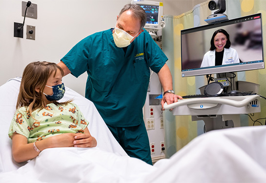 A health care provider talks to young patient at beside. A physician on telehealth interacts with them through a computer screen