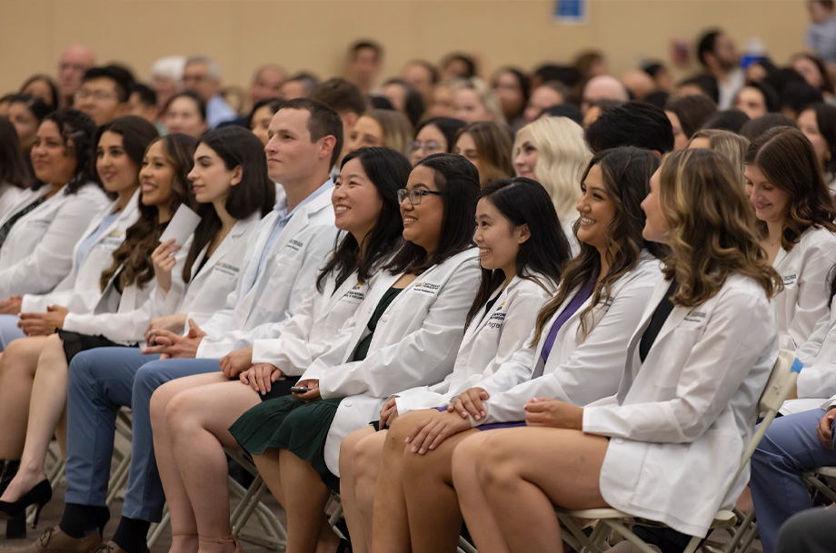 Students wearing white coats sit side-by-side in chairs during ceremony