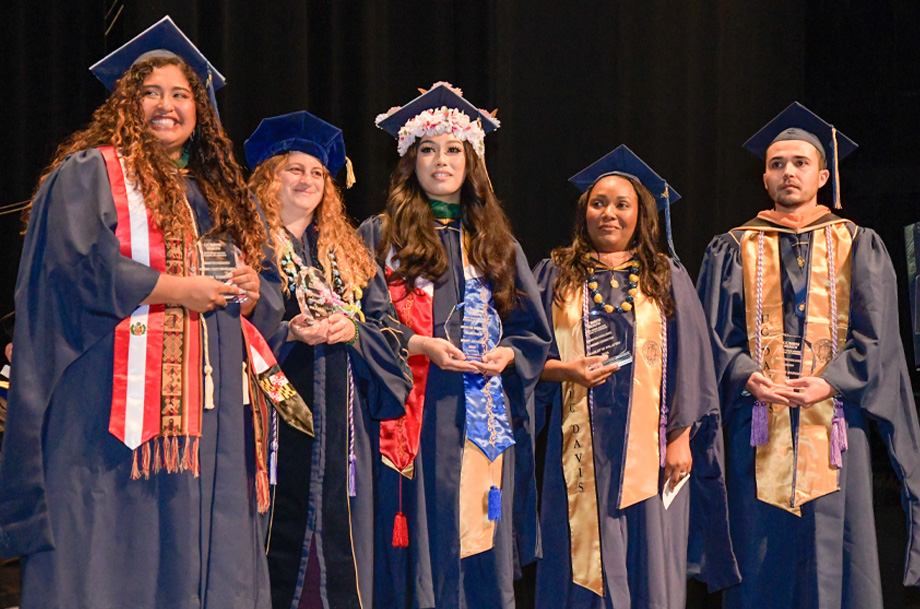 Students wearing graduation robes and holding award plaques stand side by side