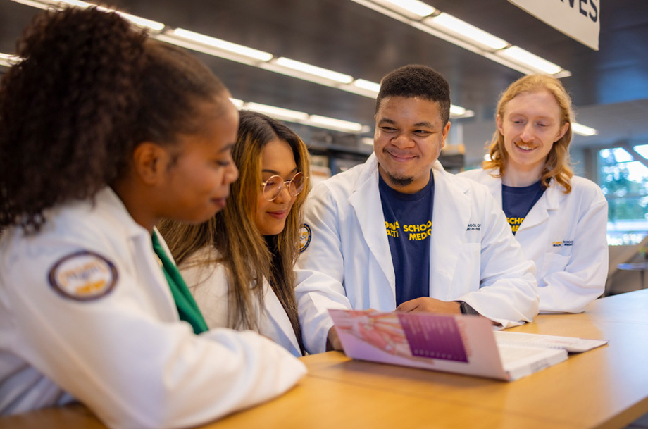 Four medical students in white coats are gathered around a table reviewing a textbook.