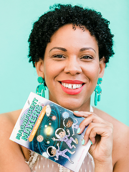 Woman with dark curly hair, green earrings and wearing a white dress holds a paperback book with the title, “Magnificent Makers.”