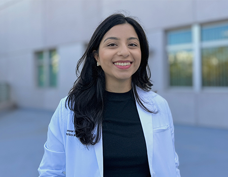 A young woman with dark hair wearing a dark shirt and white lab coat