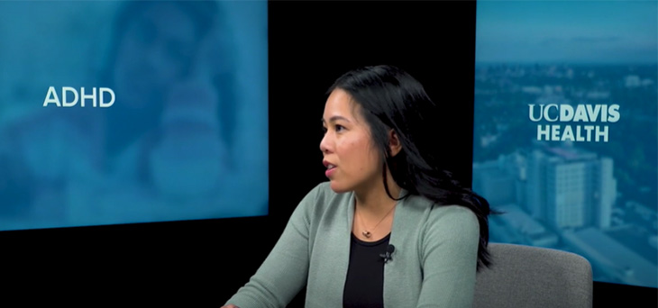 A woman with long dark hair sits at a table in front of two monitors with text “ADHD” and “UC Davis Health.”