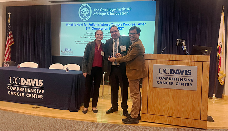 A woman and a man stand next to a man who is in the center of a group photo, holding an award.