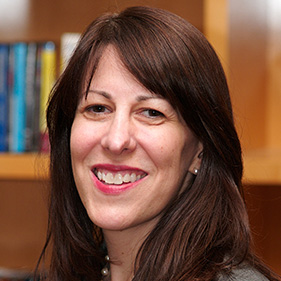 A woman with medium-length brown hair, wearing a gray suit and black shirt smiles in front of a bookcase. 