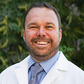 Surgeon Richard Price, wearing a white medical coat and blue shirt and tie, smiles outside. 