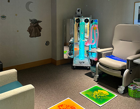 A medical office features a large, comfortable chair, colorful sensory mats on the floor, a colorful sensory station in the corner and lights on the ceiling. 