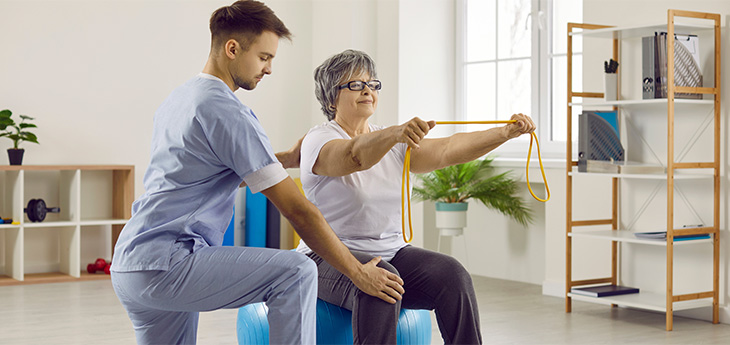 Male employee helps female senior patient with a physiotherapy exercise on fit ball. Woman on fit ball exercises with rubber band.