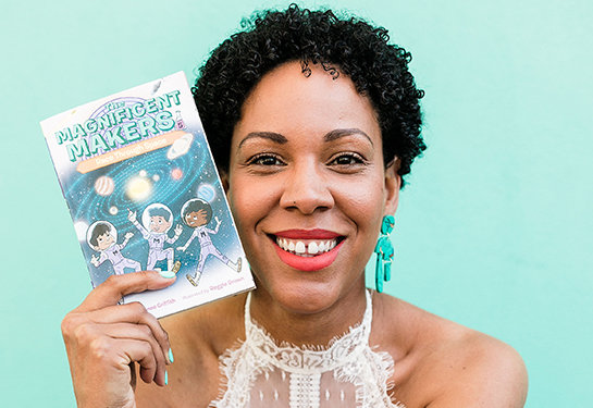 Woman with dark curly hair, green earrings and wearing a white dress holds a paperback book with the title, &#x201c;Magnificent Makers.&#x201d;